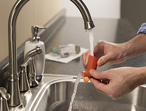 Mano que sostiene la boquilla del inhalador bajo un chorro de agua. Espaciador apoyado sobre una toalla arriba de la encimera.